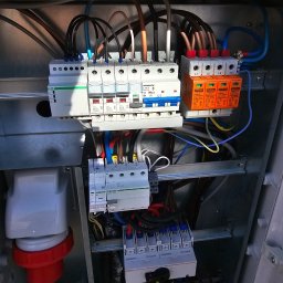 SOLSERWIS PATRYK SOŁTYSIAK - Wnętrze metalowej skrzynki rozdzielczej z zamontowanymi bezpiecznikami, ogranicznikami przepięć i przewodami o różnych kolorach, widoczne elementy instalacji elektrycznej z bliska.
