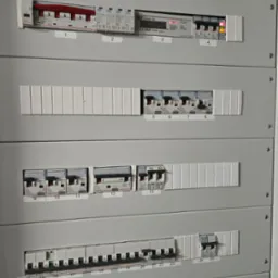 Szara rozdzielnica elektryczna z zamontowanymi bezpiecznikami i licznikiem energii, widoczne oznaczenia i etykiety na poszczególnych elementach.