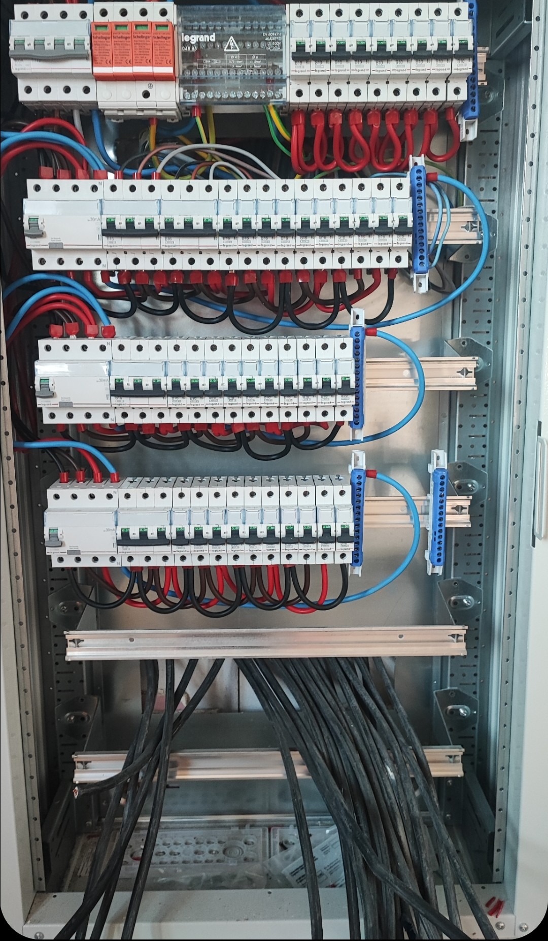 Rozdzielnica elektryczna z zabezpieczeniami przepięciowymi, licznymi wyłącznikami nadprądowymi marki Legrand, okablowaniem w kolorach czerwonym, niebieskim i czarnym, zamontowana w metalowej obudowie.