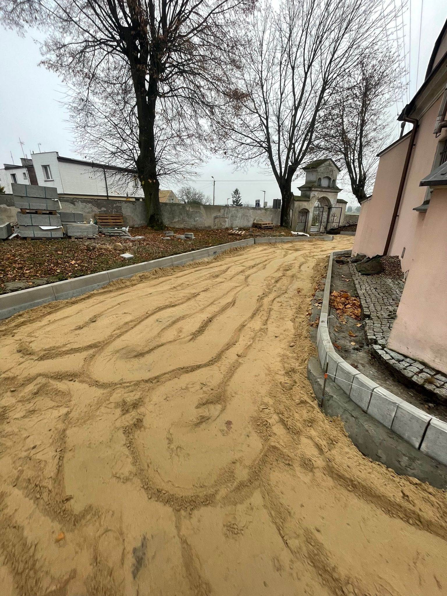 Przygotowanie podłoża z piasku pod ścieżkę z nowymi obrzeżami betonowymi, widoczne ułożone kostki obok budynku z różową elewacją i brama w tle.