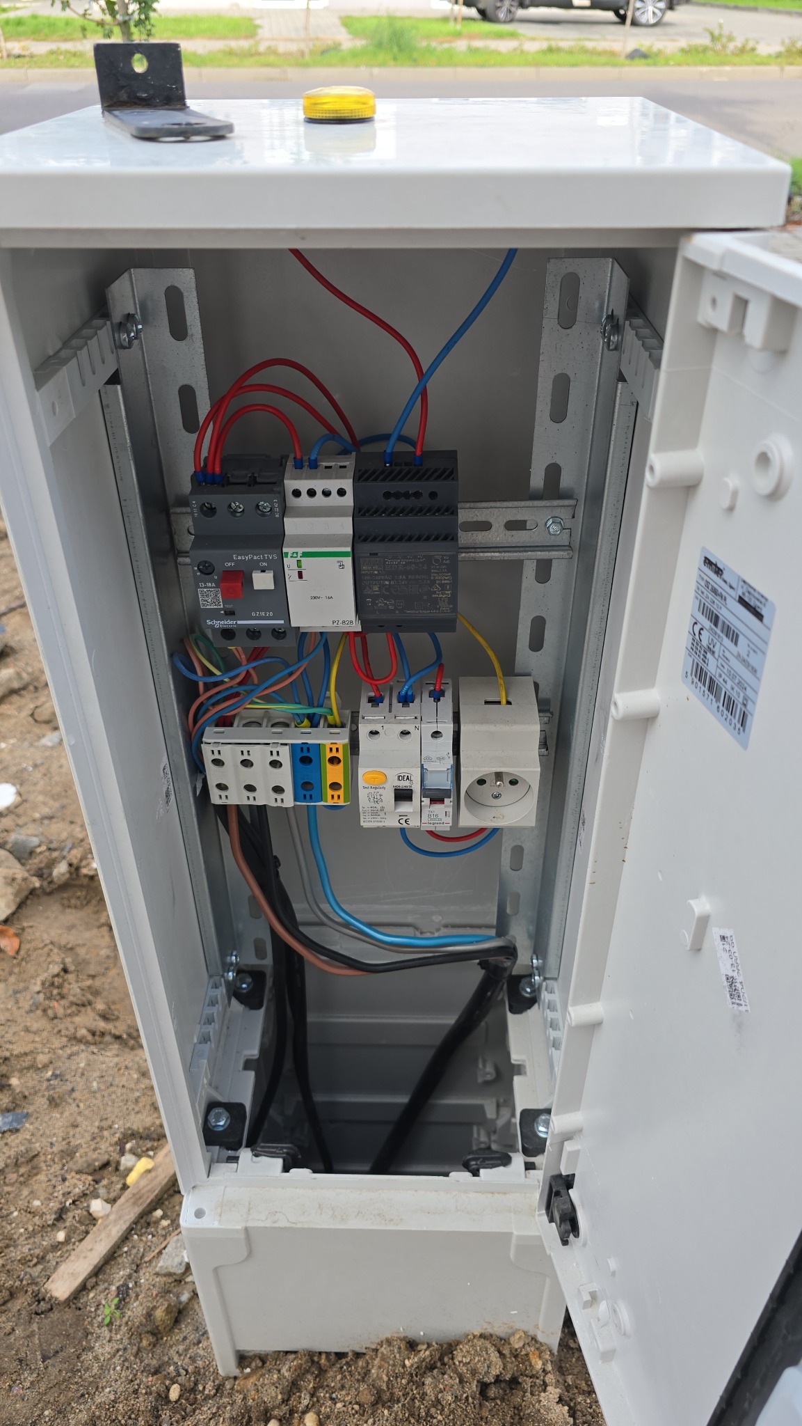 Otwarta skrzynka elektryczna na zewnątrz z zainstalowanymi bezpiecznikami, wyłącznikami, gniazdem i okablowaniem na szynie montażowej, widoczne przewody w kolorach czerwonym, niebieskim, żółtym...