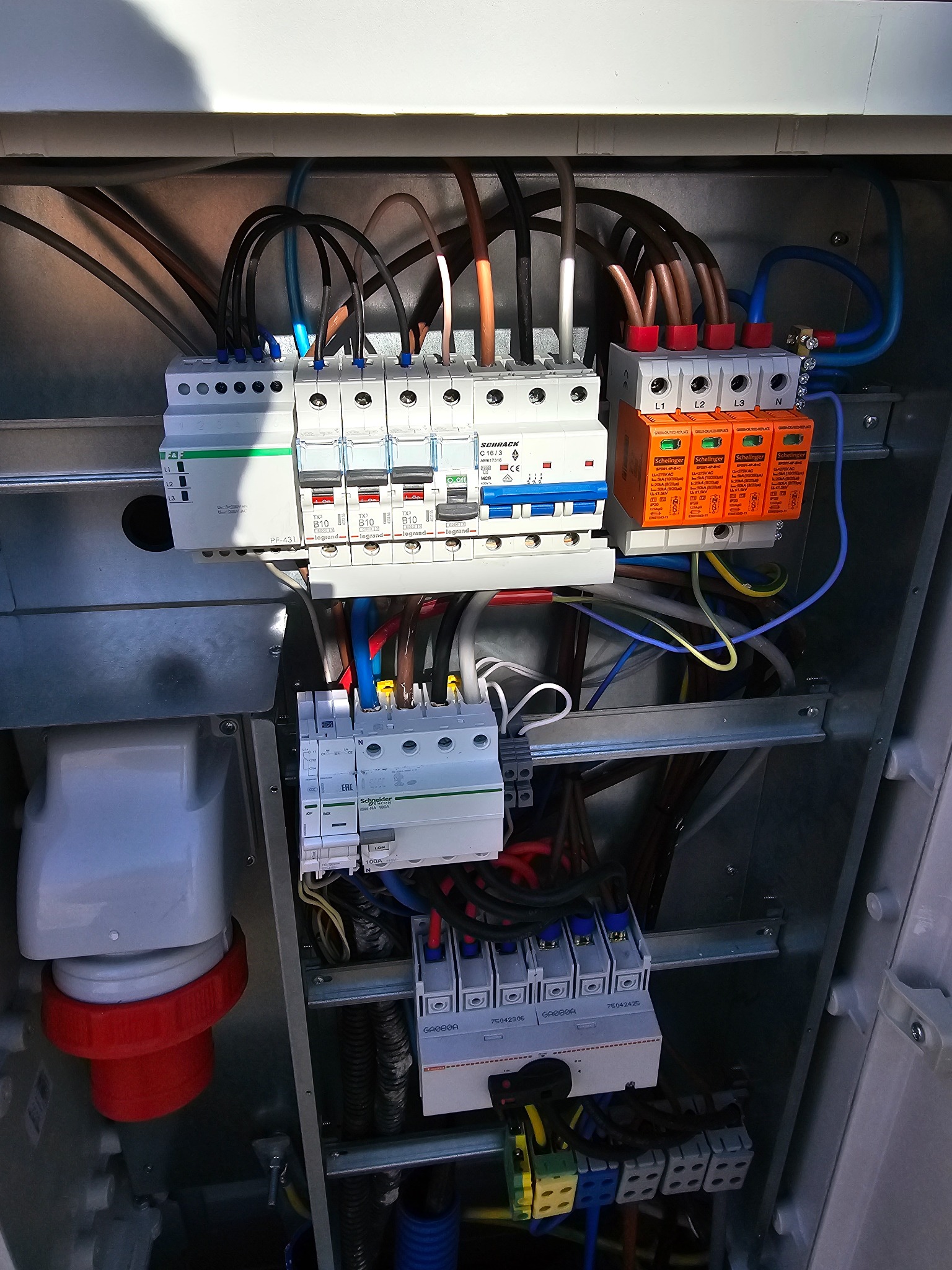 Wnętrze metalowej skrzynki rozdzielczej z zamontowanymi bezpiecznikami, ogranicznikami przepięć i przewodami o różnych kolorach, widoczne elementy instalacji elektrycznej z bliska.