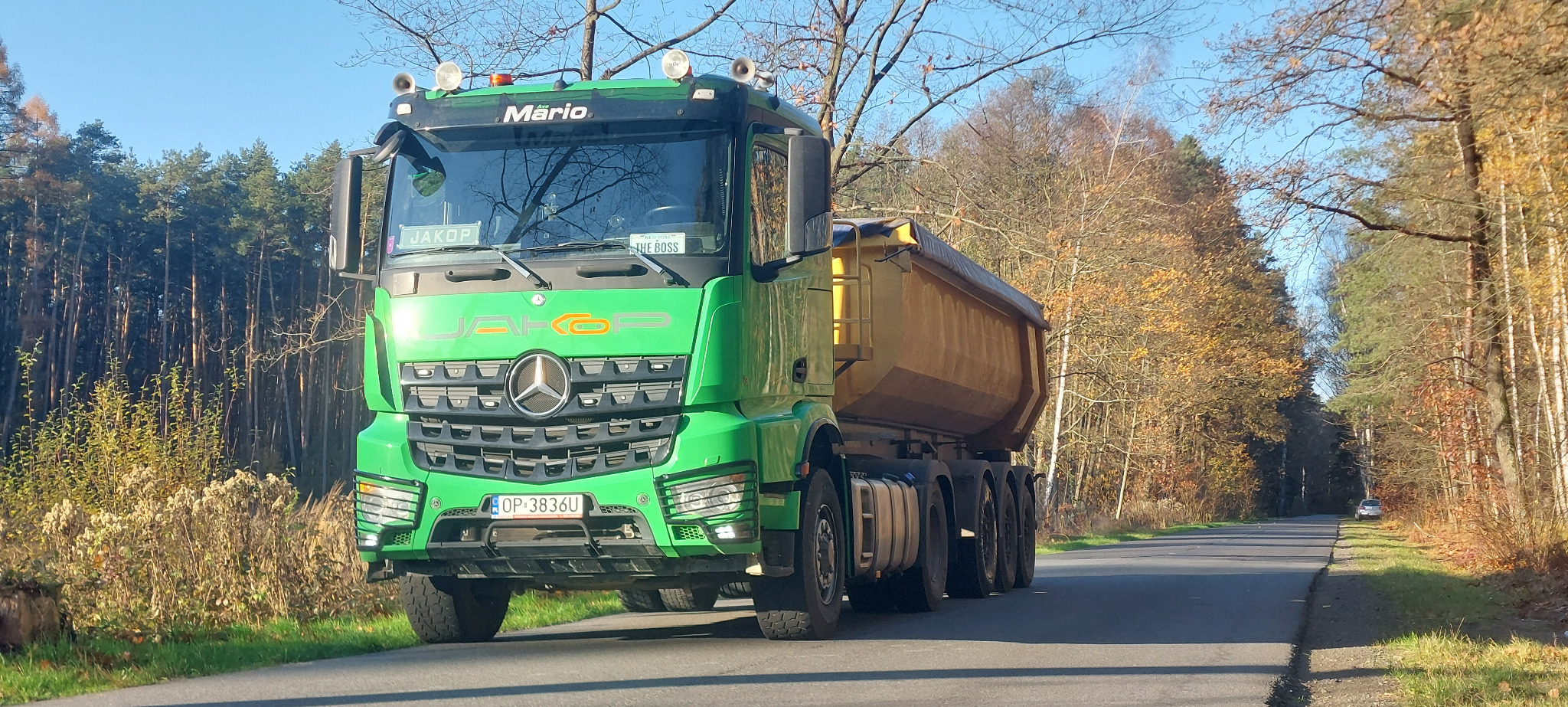Zielona ciężarówka marki Mercedes z żółtą skrzynią ładunkową stoi na asfaltowej drodze otoczonej lasem w słoneczny dzień. Na tablicy rejestracyjnej widnieje napis 'OP 3863U'.
