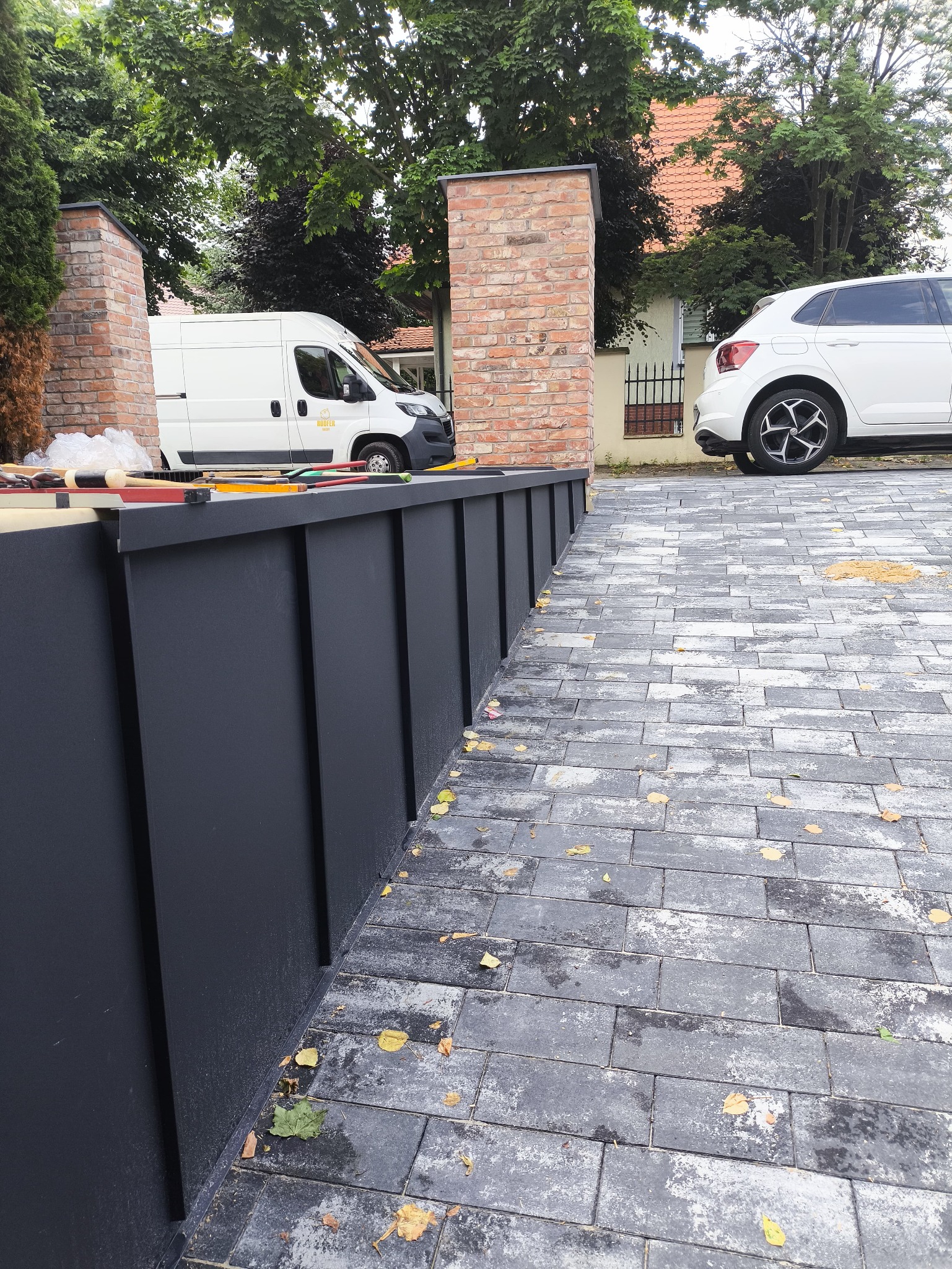 Nowoczesne obróbki blacharskie w kolorze grafitowym, z widocznymi narzędziami pracy i firmowym autem w tle. Kostka brukowa z jesiennymi liśćmi.