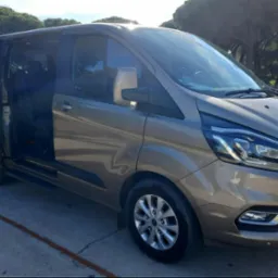 Brązowy van Ford Transit z otwartymi drzwiami przesuwnymi, zaparkowany na betonowym parkingu w słoneczny dzień.