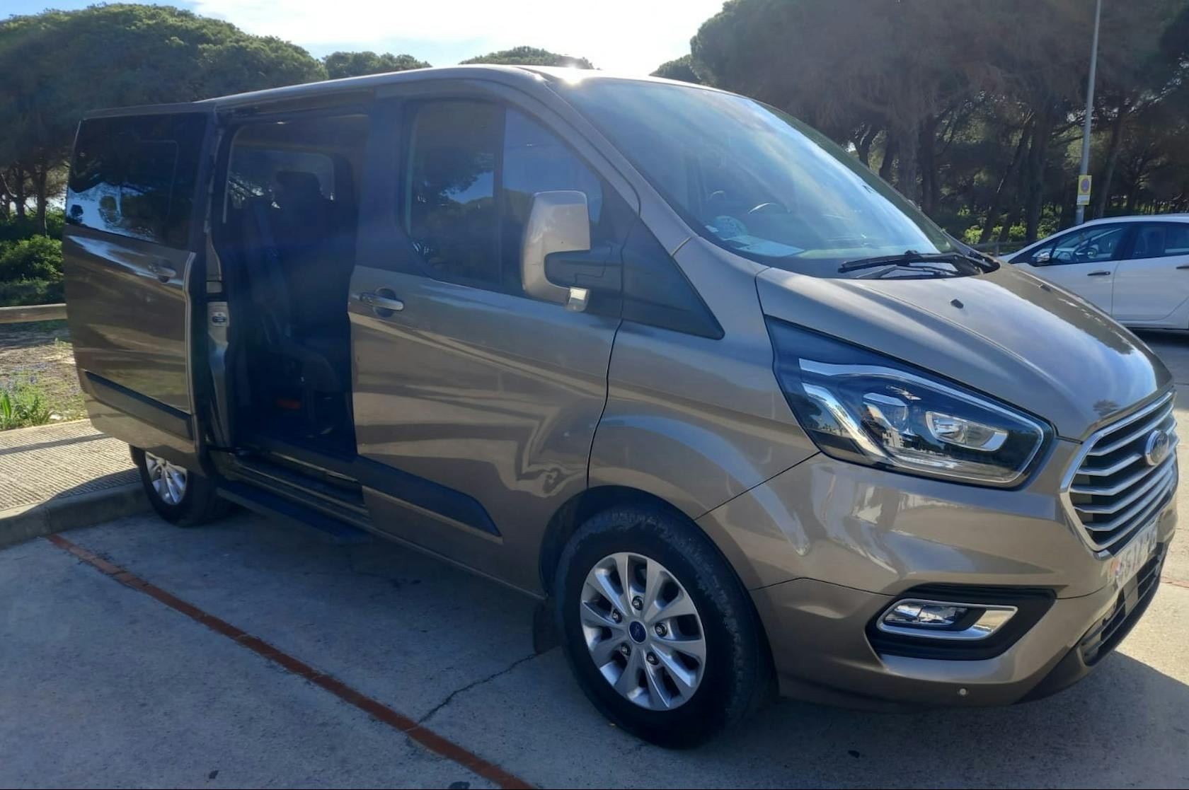 Brązowy van Ford Transit z otwartymi drzwiami przesuwnymi, zaparkowany na betonowym parkingu w słoneczny dzień.