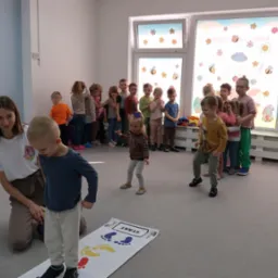 Animator prowadzi zajęcia ruchowe z dziećmi w przedszkolu, wykorzystując matę z kolorowymi odciskami stóp, na tle grupy dzieci i okna z dekoracjami.