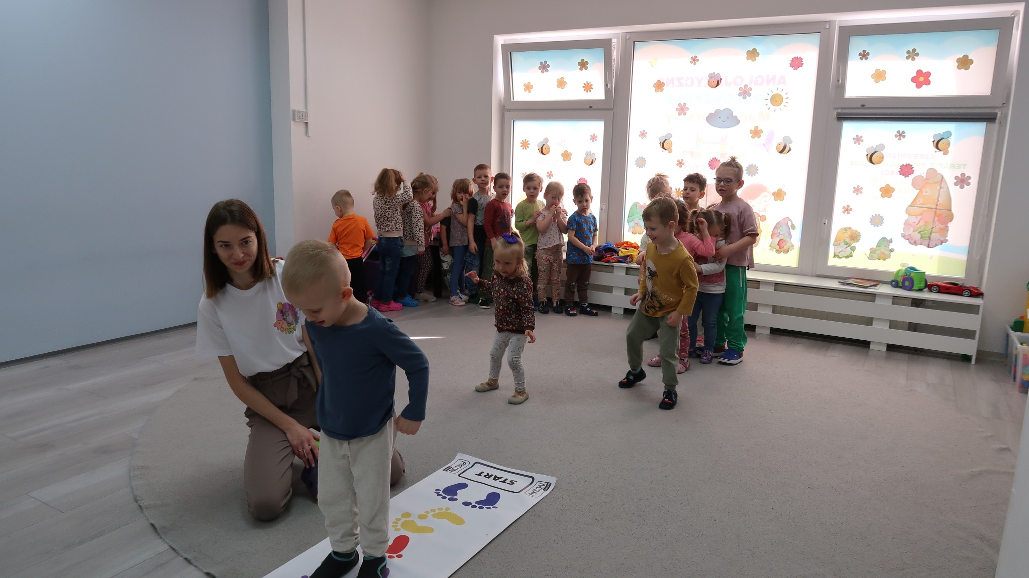 Animator prowadzi zajęcia ruchowe z dziećmi w przedszkolu, wykorzystując matę z kolorowymi odciskami stóp, na tle grupy dzieci i okna z dekoracjami.