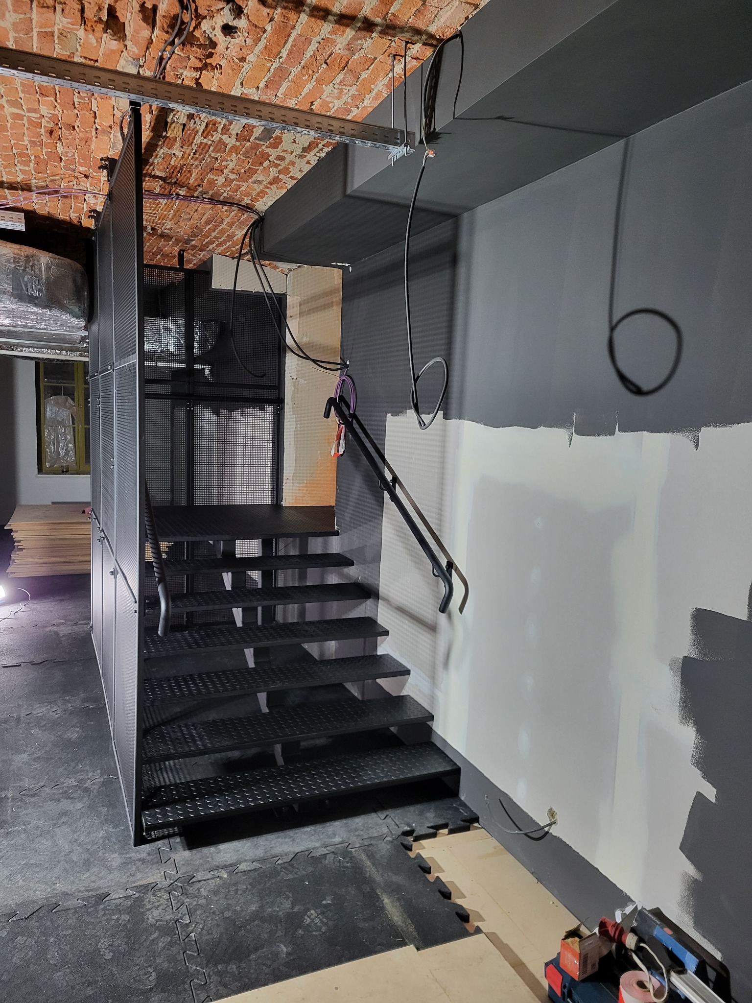 Czarne schody z metalu z perforowaną obudową w surowym wnętrzu z ceglanym stropem i szarą ścianą w trakcie malowania. Widoczne narzędzia i materiały budowlane.