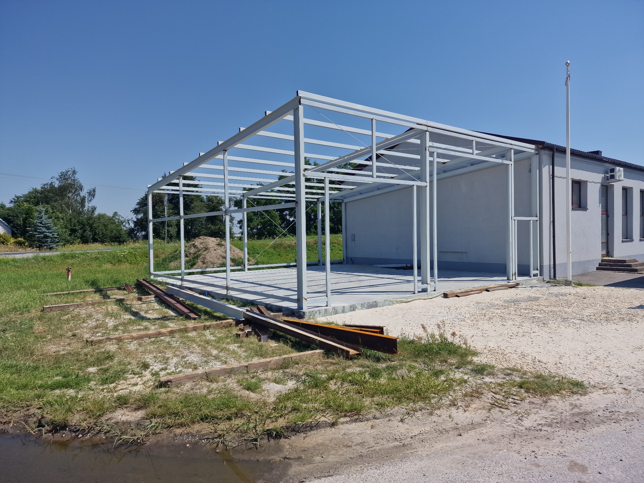 Konstrukcja stalowa hali w trakcie budowy, widok z zewnątrz. Białe profile tworzą szkielet budynku na tle błękitnego nieba i budynku administracyjnego.