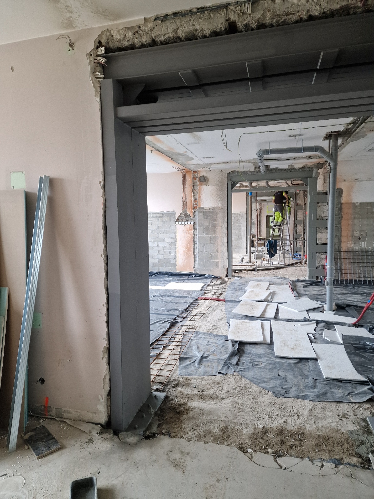 Montaż stalowej konstrukcji w surowym wnętrzu. Widoczne elementy nośne, instalacje i przygotowania do dalszych prac wykończeniowych. W tle pracownik na drabinie.