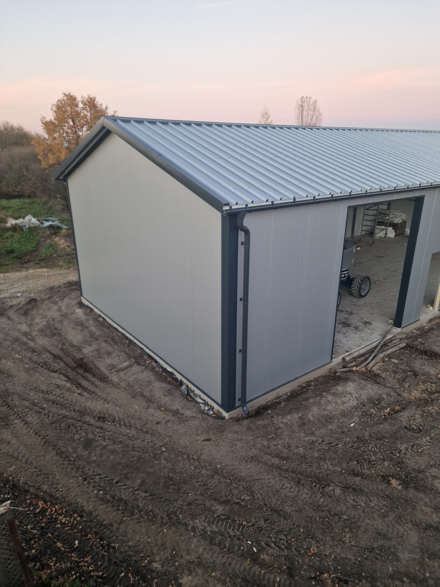 Szara hala z blachy trapezowej w trakcie budowy, widok z zewnątrz. W tle drzewa i niebo o zmierzchu. Na podłożu ślady opon.