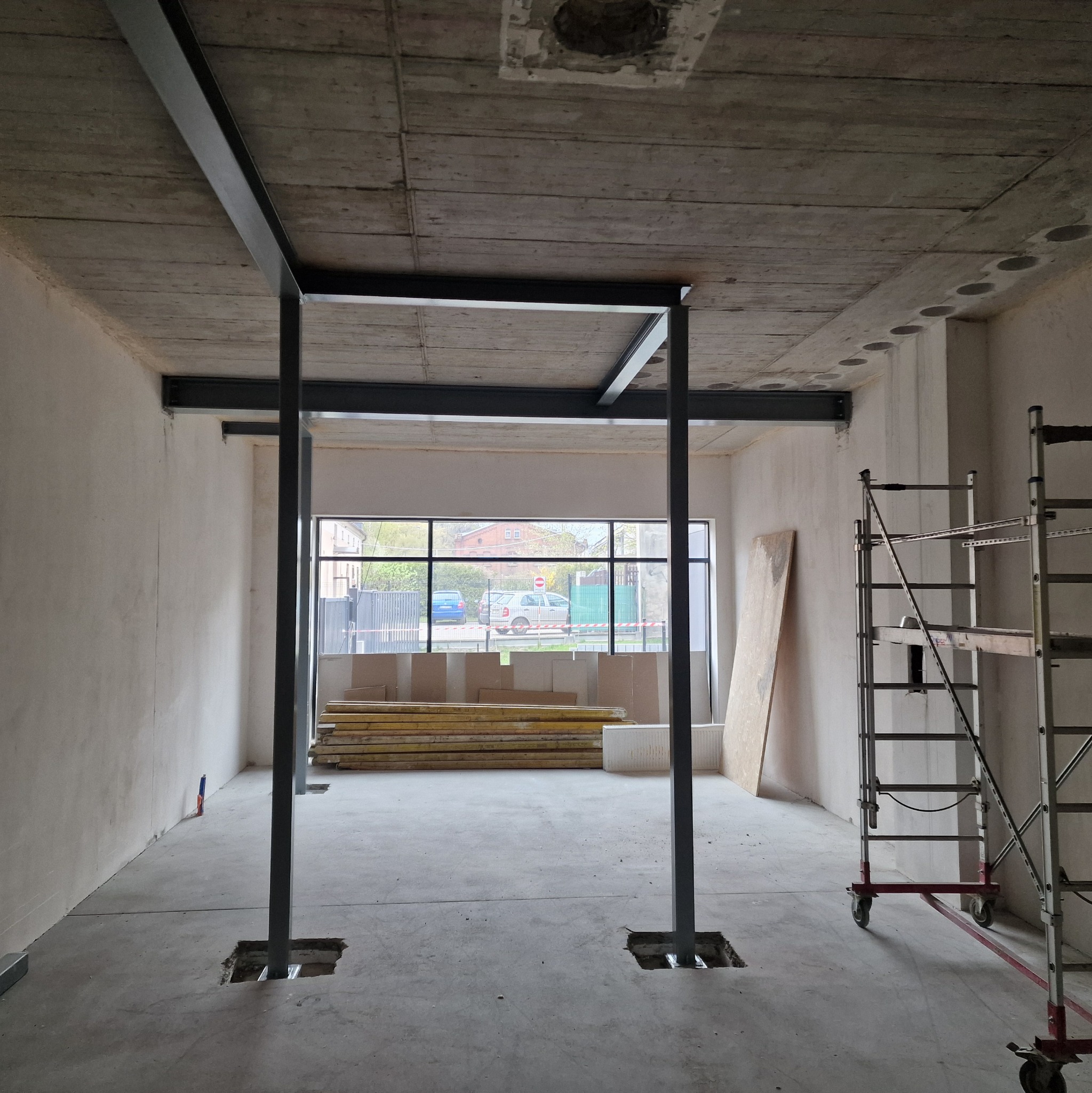Wnętrze w trakcie budowy hali z widoczną stalową konstrukcją nośną, surowym betonowym stropem i ścianami. W tle okno z widokiem na ulicę i rusztowanie.