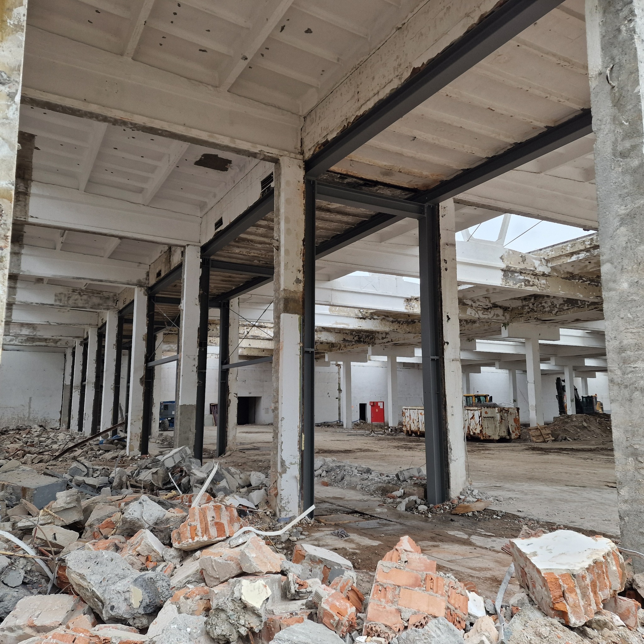 Wnętrze hali w trakcie budowy z widocznymi elementami konstrukcji stalowej i gruzem. Widoczne słupy i belki, a w tle sprzęt budowlany.