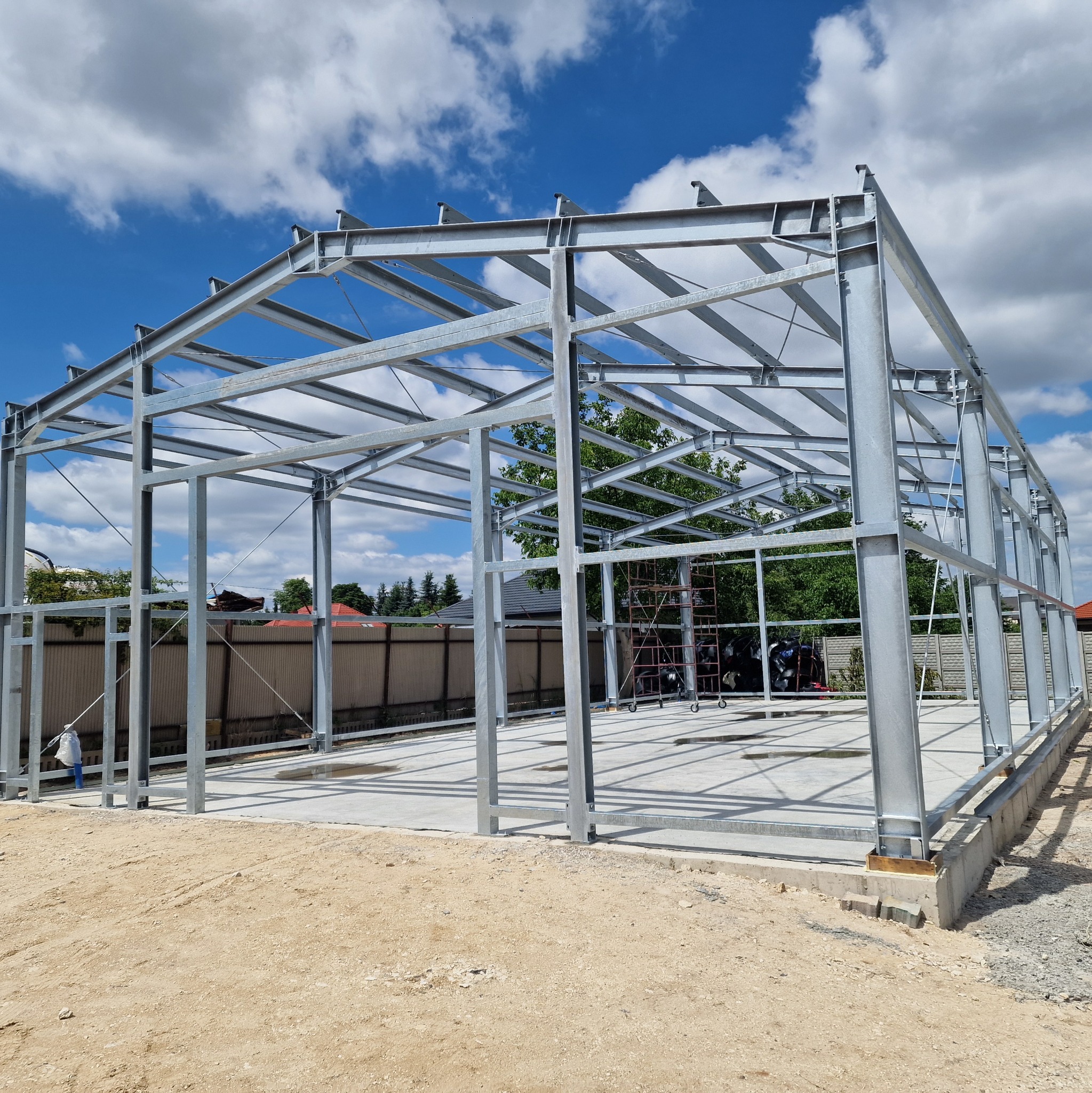 Konstrukcja stalowa hali w trakcie budowy, widoczne stalowe ramy i słupy na betonowej podstawie, błękitne niebo z chmurami w tle. Częściowo widoczne ogrodzenie i zieleń.