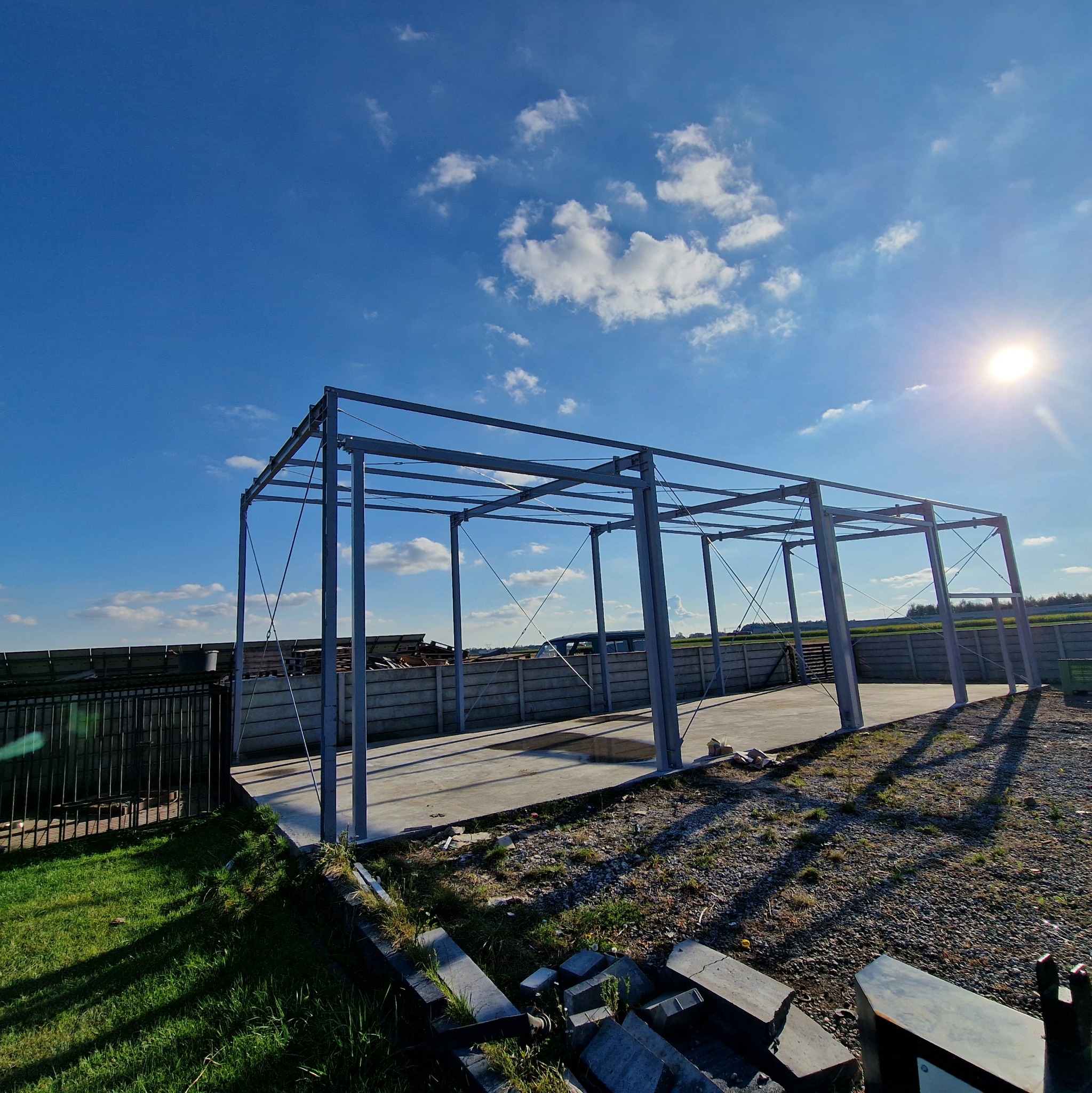 Stalowa konstrukcja hali w trakcie budowy, widok z dołu na błękitne niebo z chmurami. Słoneczny dzień, betonowa podstawa, widoczne elementy mocujące.