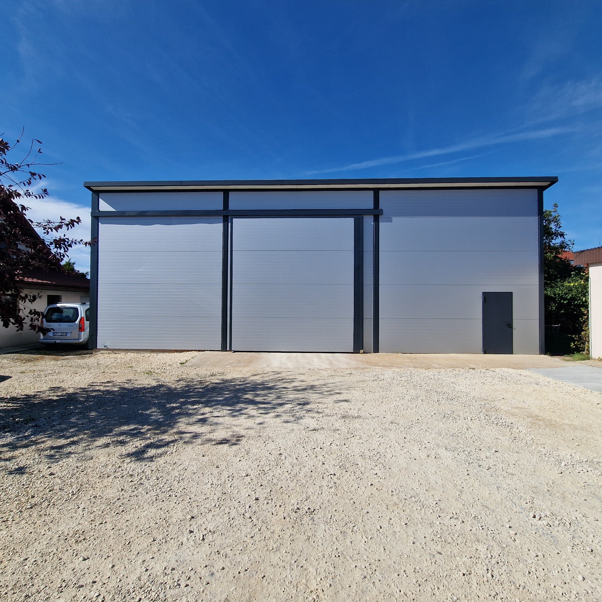 Nowoczesna hala magazynowa z szarej blachy trapezowej, z przesuwną bramą i drzwiami wejściowymi. Błękitne niebo w tle, żwirowe podłoże.