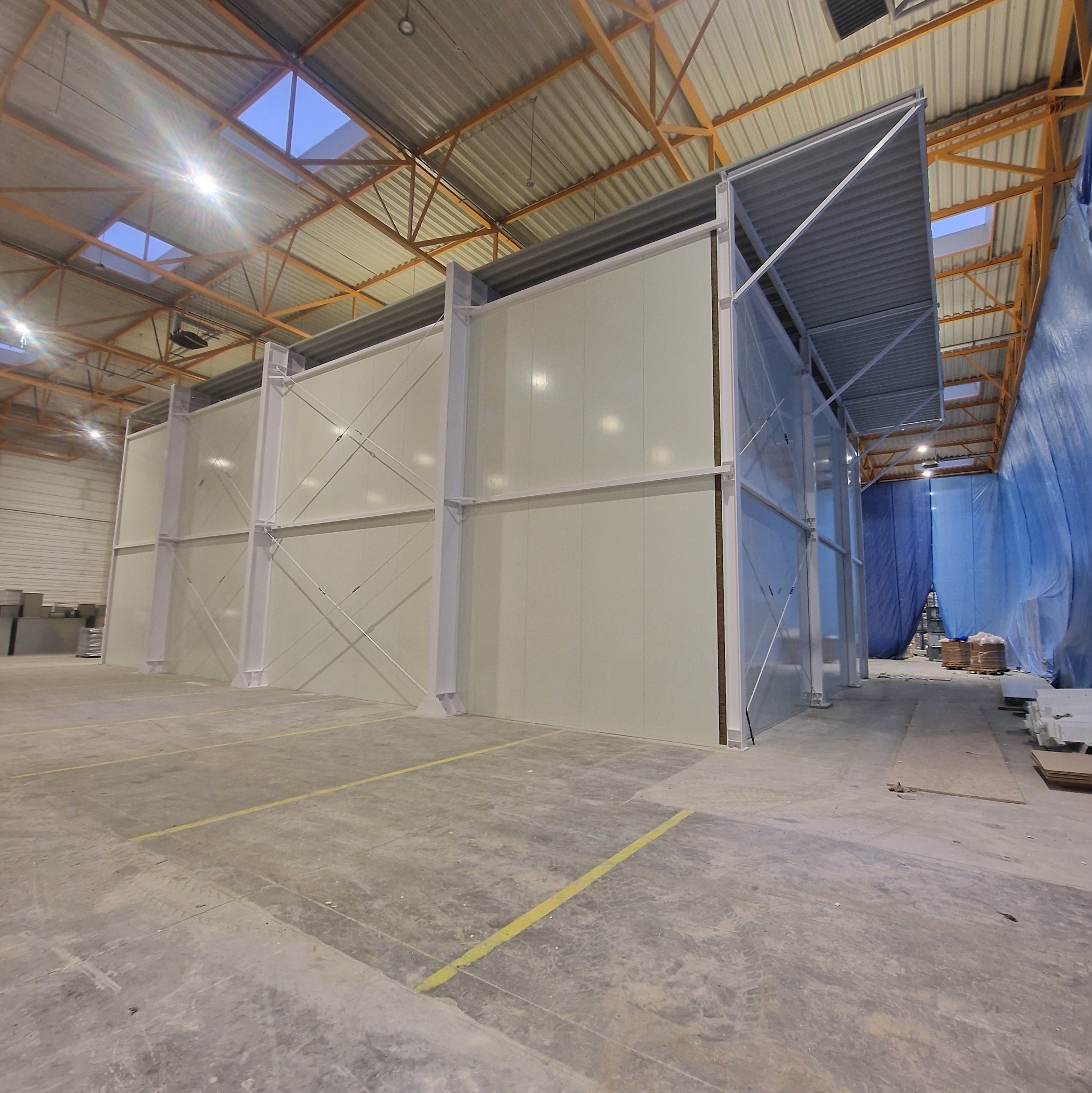 Wnętrze hali produkcyjnej z widocznymi ścianami działowymi o konstrukcji stalowej i panelach. Widoczne elementy konstrukcji dachu i oświetlenie.