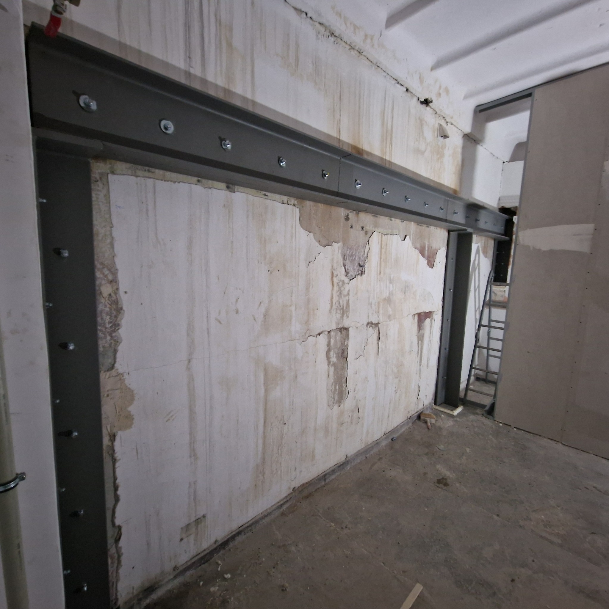 Wzmocnienie ściany nośnej stalową konstrukcją skręcaną śrubami. Widoczne ślady po usuniętych fragmentach tynku. W tle drabina.