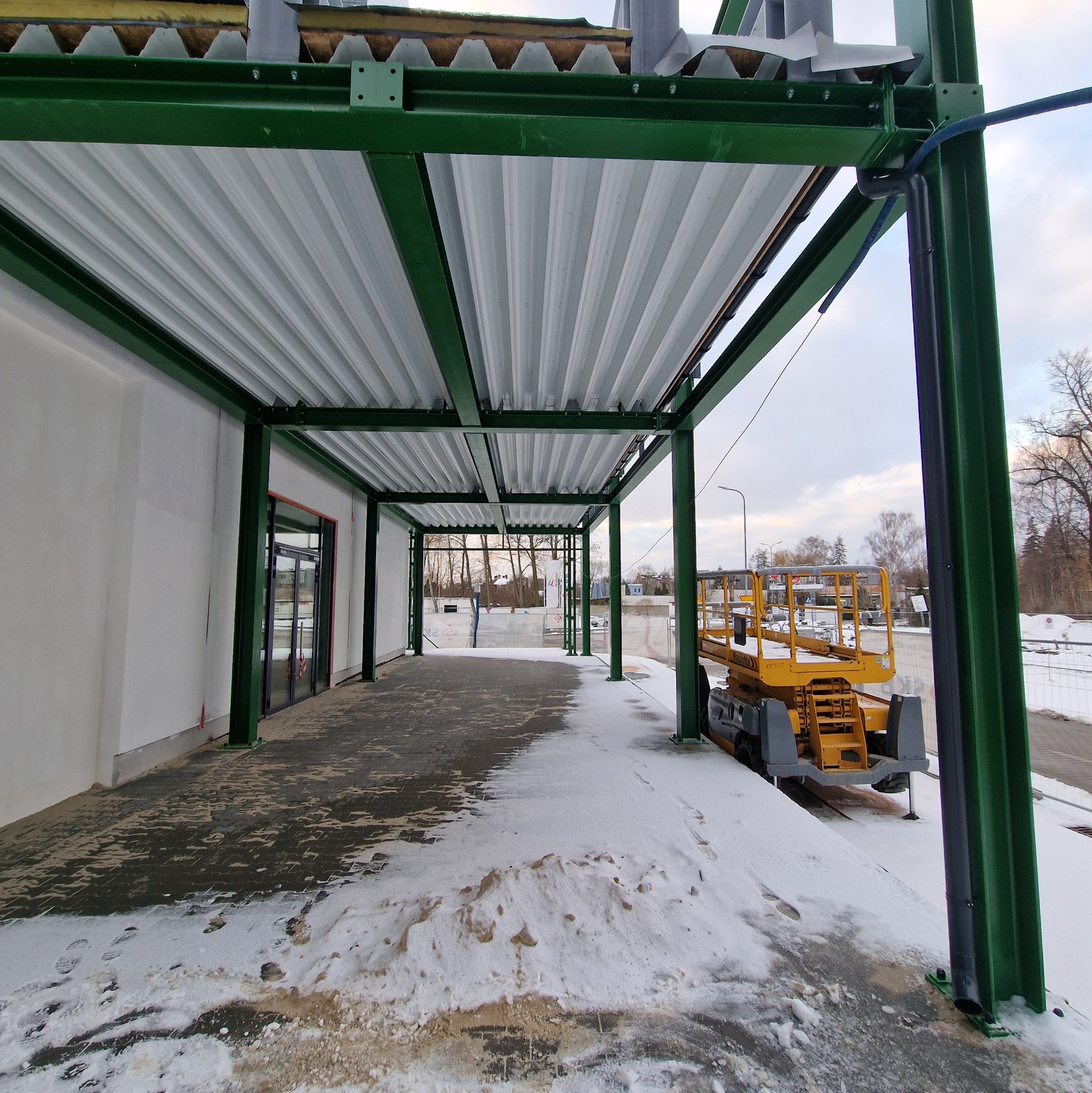 Konstrukcja stalowa hali w Karlin z zielonymi słupami i białym dachem z blachy trapezowej. Na zewnątrz żółty podnośnik koszowy i śnieg.