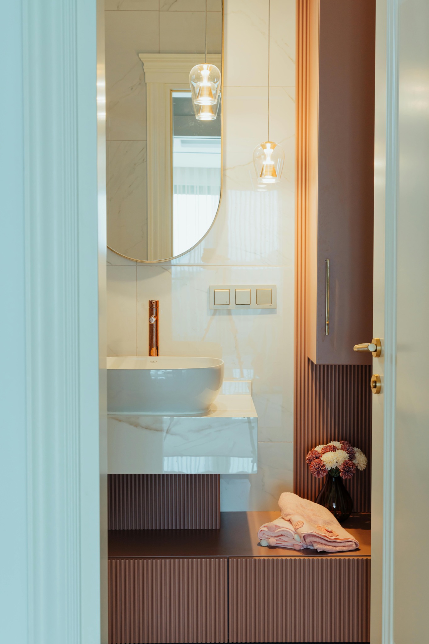 Elegancka łazienka z umywalką nablatową, okrągłym lustrem w złotej ramie, złotą armaturą i wiszącymi lampami, wykończona w bieli i odcieniach różu z dekoracyjnymi panelami ściennymi i kwiatami...