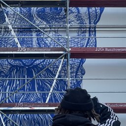 Krzysztof Czarnecki - Mural w trakcie tworzenia na rusztowaniu, widok zza pleców osoby w czarnej kurtce z trzema białymi paskami na rękawach, wzór na ścianie w odcieniach błękitu i bieli.