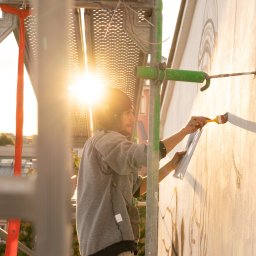 Krzysztof Czarnecki - Artysta na rusztowaniu maluje mural na ścianie budynku w promieniach zachodzącego słońca, używając pędzla i wzoru z kartki.