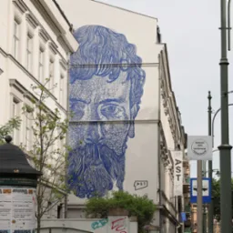 Niebieski mural przedstawiający twarz brodatego mężczyzny na bocznej ścianie kamienicy, widok z ulicy z elementami zieleni i słupem ogłoszeniowym.
