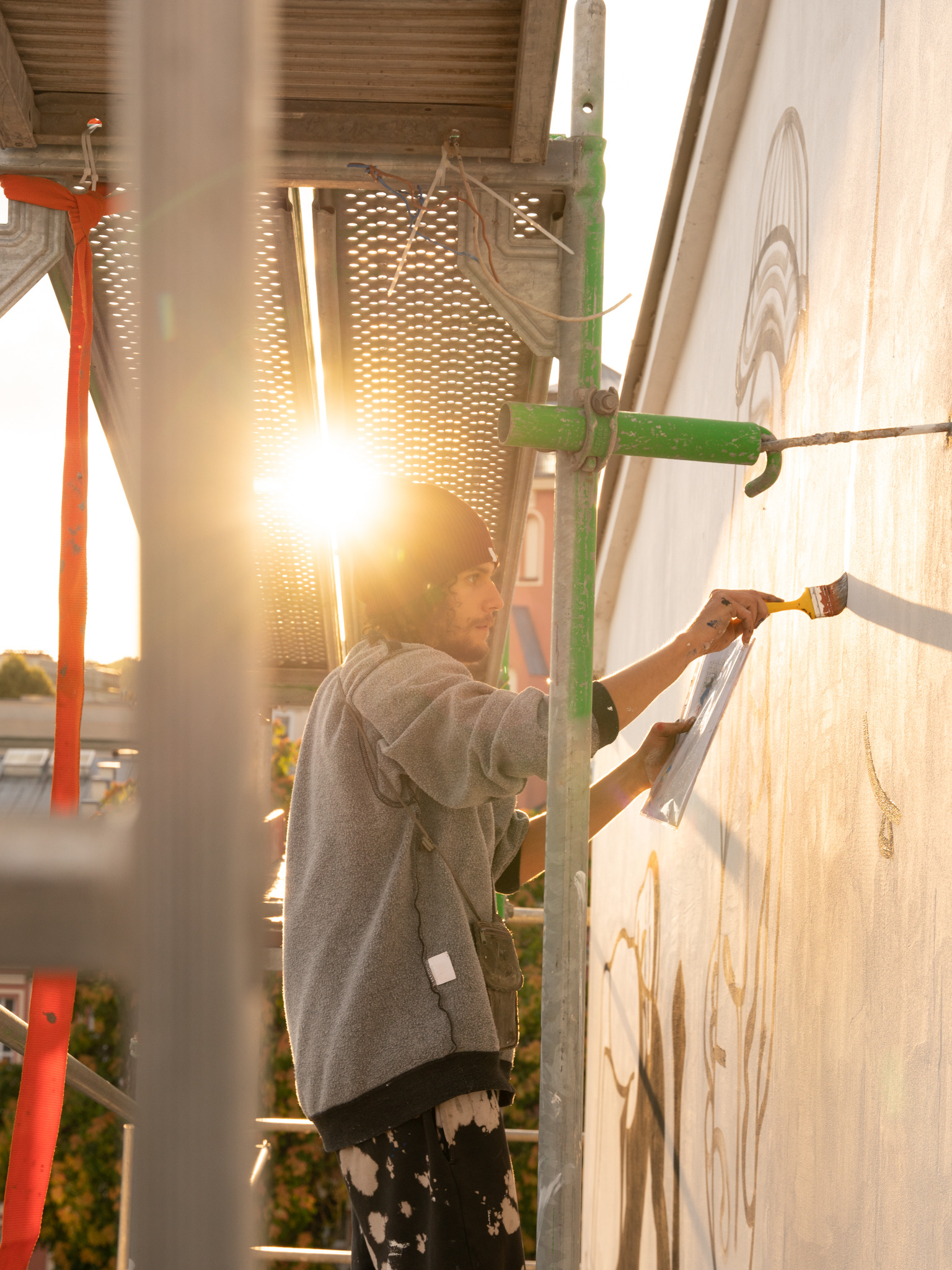 Artysta na rusztowaniu maluje mural na ścianie budynku w promieniach zachodzącego słońca, używając pędzla i wzoru z kartki.