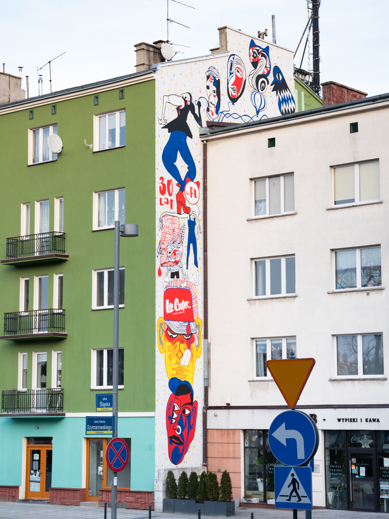 Kolorowy mural na ścianie budynku przedstawiający postacie i abstrakcyjne formy, obok budynek z zieloną fasadą i balkonikami.