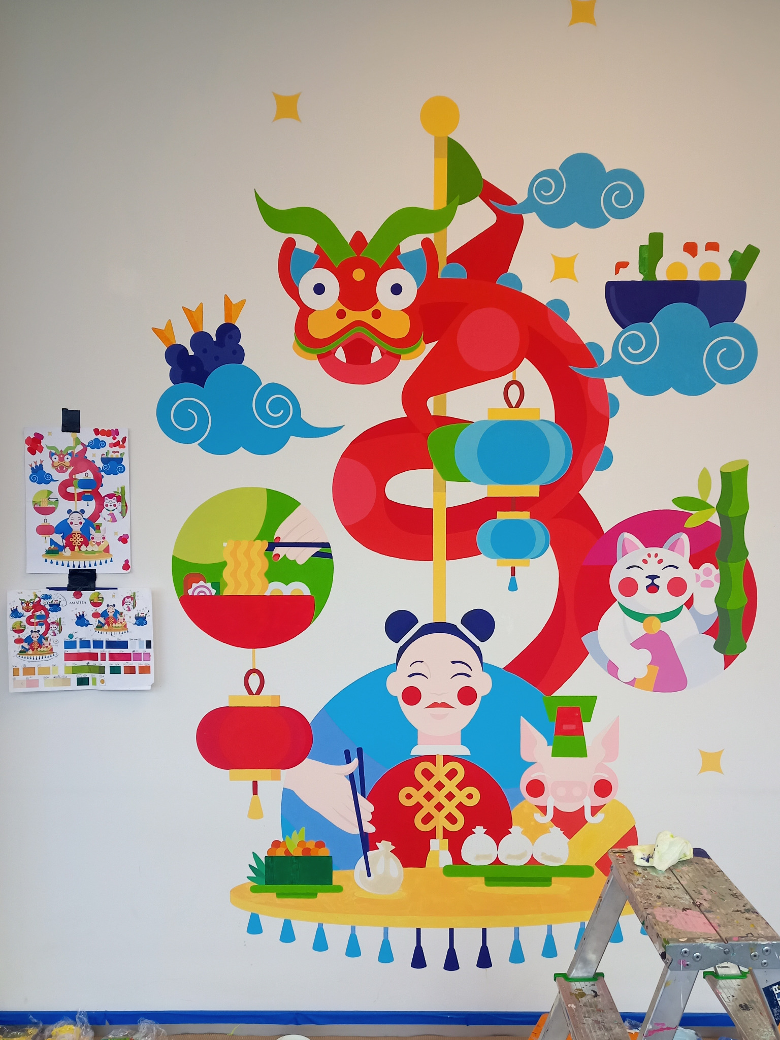 Ściana z kolorową, płaską ilustracją w stylu azjatyckim przedstawiającą smoka, kota machającego łapką, postacie i potrawy, z drabinką malarską w prawym dolnym rogu.