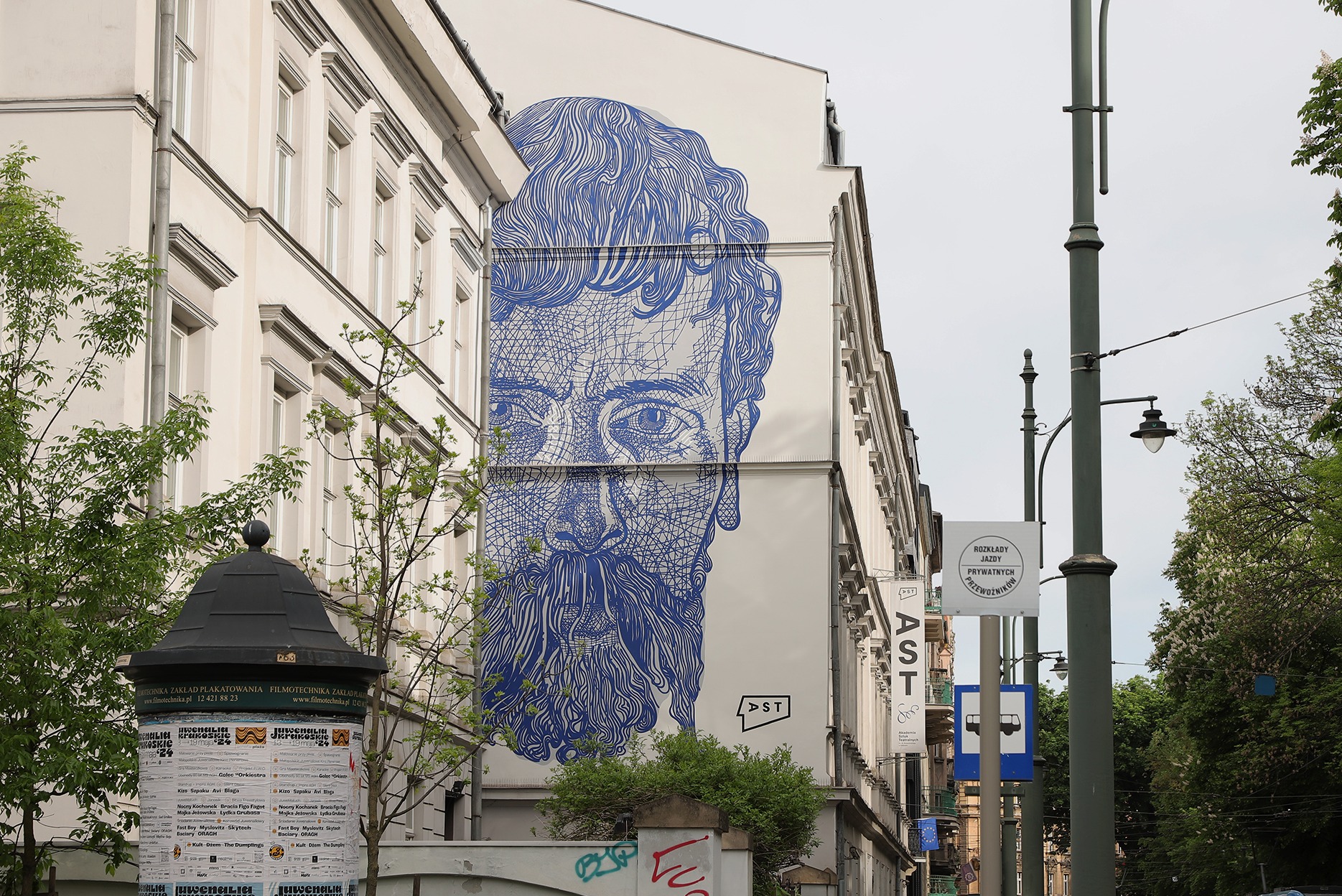 Niebieski mural przedstawiający twarz brodatego mężczyzny na bocznej ścianie kamienicy, widok z ulicy z elementami zieleni i słupem ogłoszeniowym.