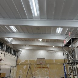 DDD OZON Michał Zięba - Wnętrze sali gimnastycznej w trakcie prac remontowych: pomalowany sufit z oświetleniem rastrowym, częściowo odnowione ściany z koszami do koszykówki, rusztowanie z plandeką ochronną, drabina oparta...