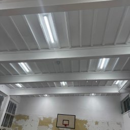DDD OZON Michał Zięba - Wnętrze sali gimnastycznej w trakcie odnawiania: pomalowany sufit z oświetleniem, ściany częściowo przygotowane do malowania, widoczny kosz do koszykówki.