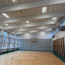 DDD OZON Michał Zięba - Wnętrze sali gimnastycznej z parkietem w odcieniach beżu, koszem do koszykówki na szarej ścianie i rzędem okien po lewej stronie, oświetlone białymi lampami sufitowymi.