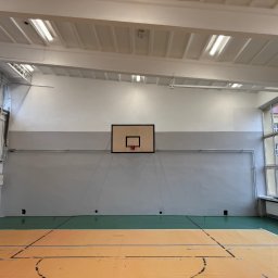 DDD OZON Michał Zięba - Wnętrze sali gimnastycznej z parkietem, drabinkami gimnastycznymi, koszem do koszykówki i oknami z widokiem na budynek na zewnątrz, świeżo odmalowane ściany w dwóch odcieniach szarości.