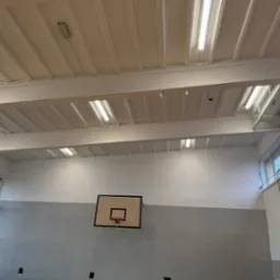 Wnętrze sali gimnastycznej po odświeżeniu: pomalowany sufit z oświetleniem rastrowym, ściany dwukolorowe (szary dół, biała góra), kosz do koszykówki na ścianie.