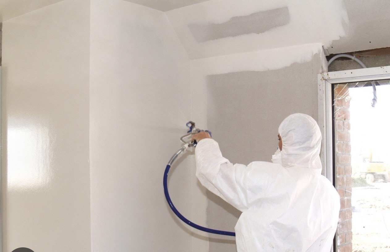 Pracownik w kombinezonie maluje ścianę natryskowo na biało, widoczne świeżo pomalowane fragmenty i niepomalowane obszary oraz niezagruntowany sufit, niebieski wąż od pistoletu natryskowego.