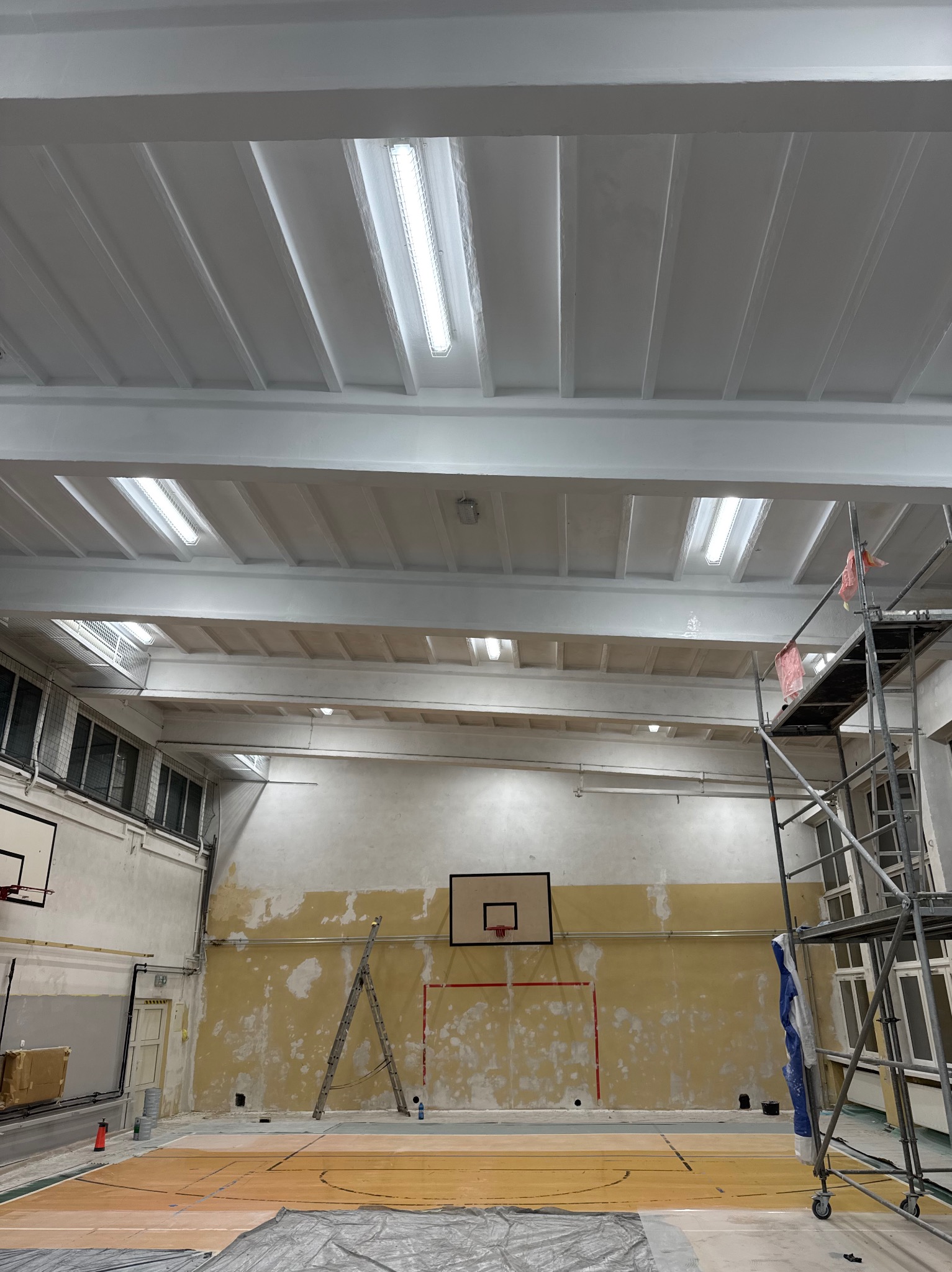 Wnętrze sali gimnastycznej w trakcie prac remontowych: pomalowany sufit z oświetleniem rastrowym, częściowo odnowione ściany z koszami do koszykówki, rusztowanie z plandeką ochronną, drabina oparta...