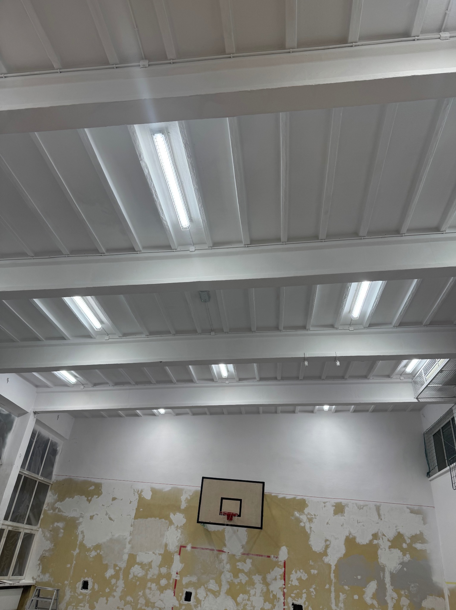 Wnętrze sali gimnastycznej w trakcie odnawiania: pomalowany sufit z oświetleniem, ściany częściowo przygotowane do malowania, widoczny kosz do koszykówki.