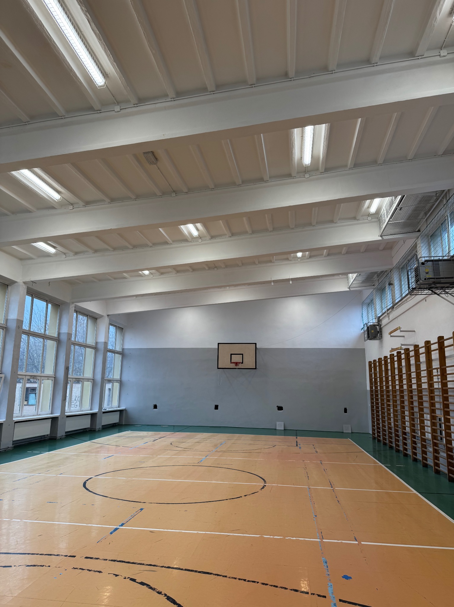 Wnętrze sali gimnastycznej z parkietem w odcieniach beżu, koszem do koszykówki na szarej ścianie i rzędem okien po lewej stronie, oświetlone białymi lampami sufitowymi.