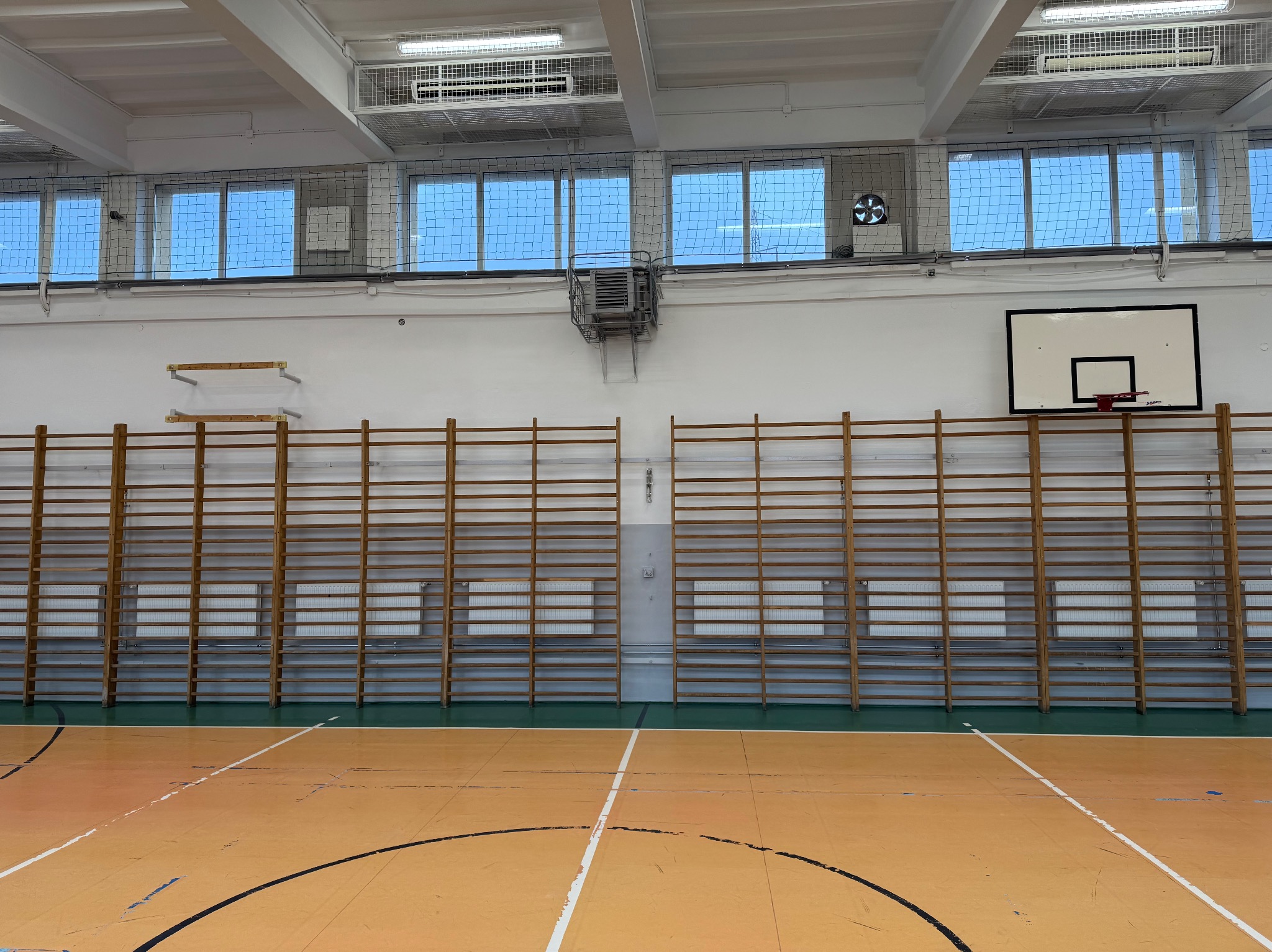 Wnętrze sali gimnastycznej z drabinkami gimnastycznymi, koszem do koszykówki i klimatyzatorem pod sufitem.