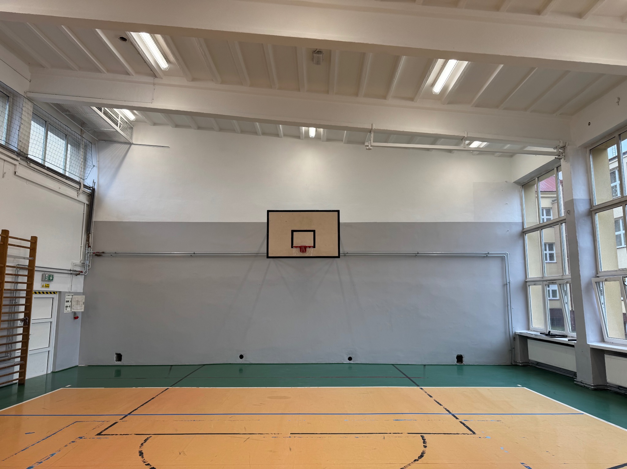 Wnętrze sali gimnastycznej z parkietem, drabinkami gimnastycznymi, koszem do koszykówki i oknami z widokiem na budynek na zewnątrz, świeżo odmalowane ściany w dwóch odcieniach szarości.