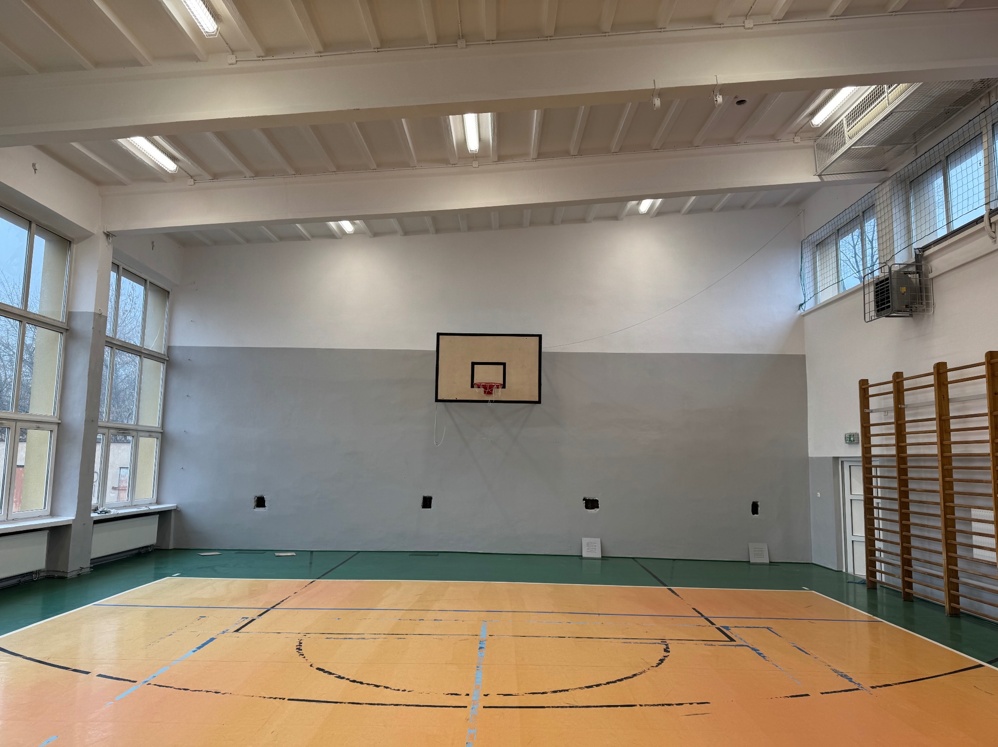 Wnętrze sali gimnastycznej z parkietem w odcieniach pomarańczy i zieleni, koszem do koszykówki na szarej ścianie, drabinkami gimnastycznymi oraz oknami z widokiem na drzewa.