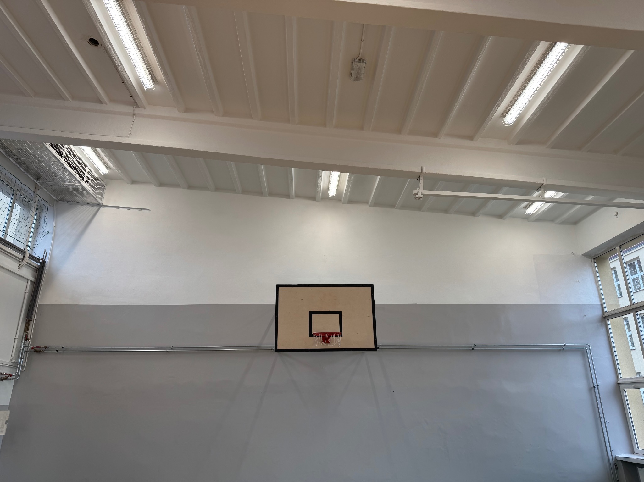 Wnętrze sali gimnastycznej po malowaniu: ściany pomalowane na szaro i biało, widoczny kosz do koszykówki, rury instalacyjne i oświetlenie sufitowe.