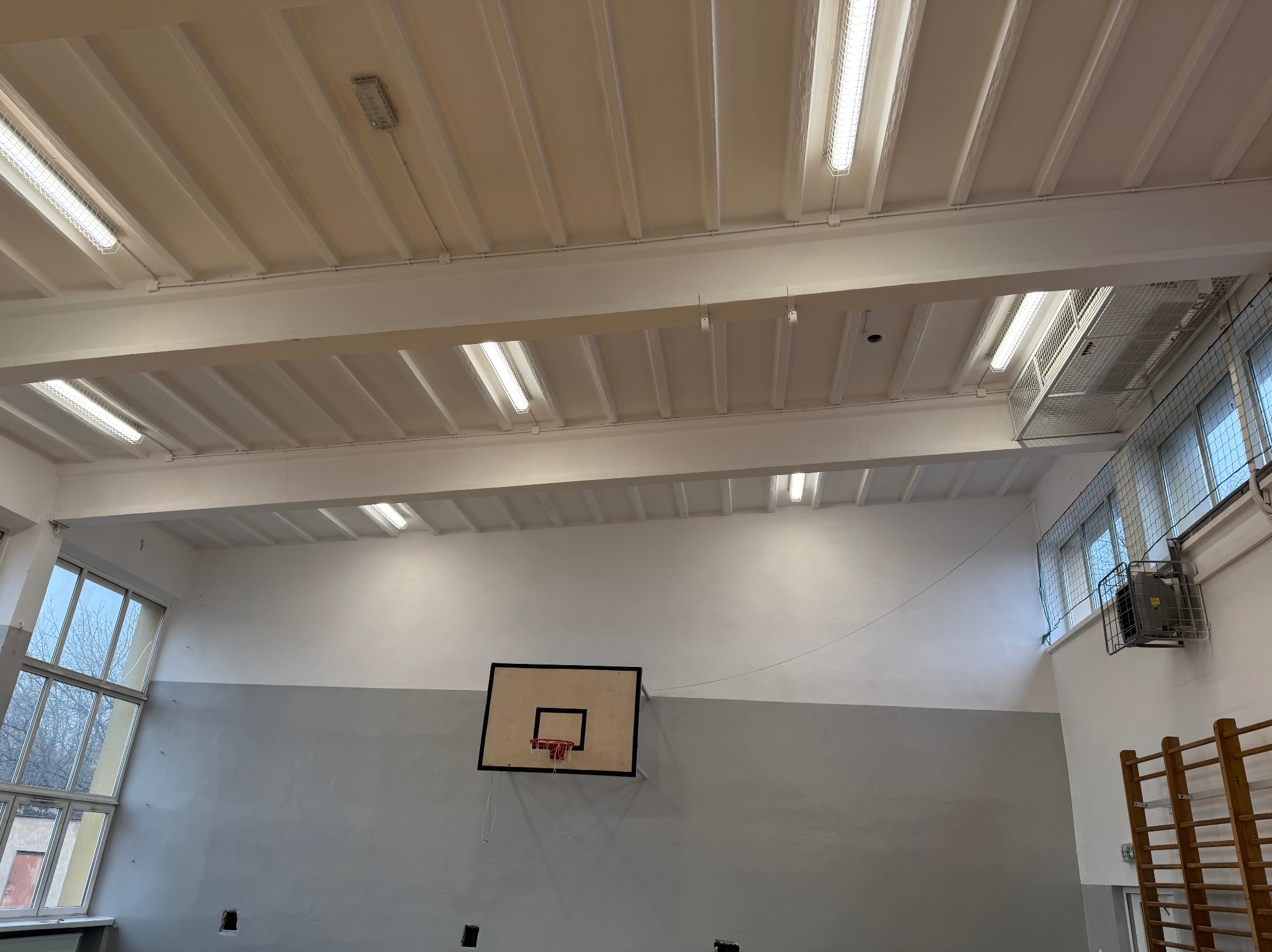 Wnętrze sali gimnastycznej po odświeżeniu: pomalowany sufit z oświetleniem rastrowym, ściany dwukolorowe (szary dół, biała góra), kosz do koszykówki na ścianie.