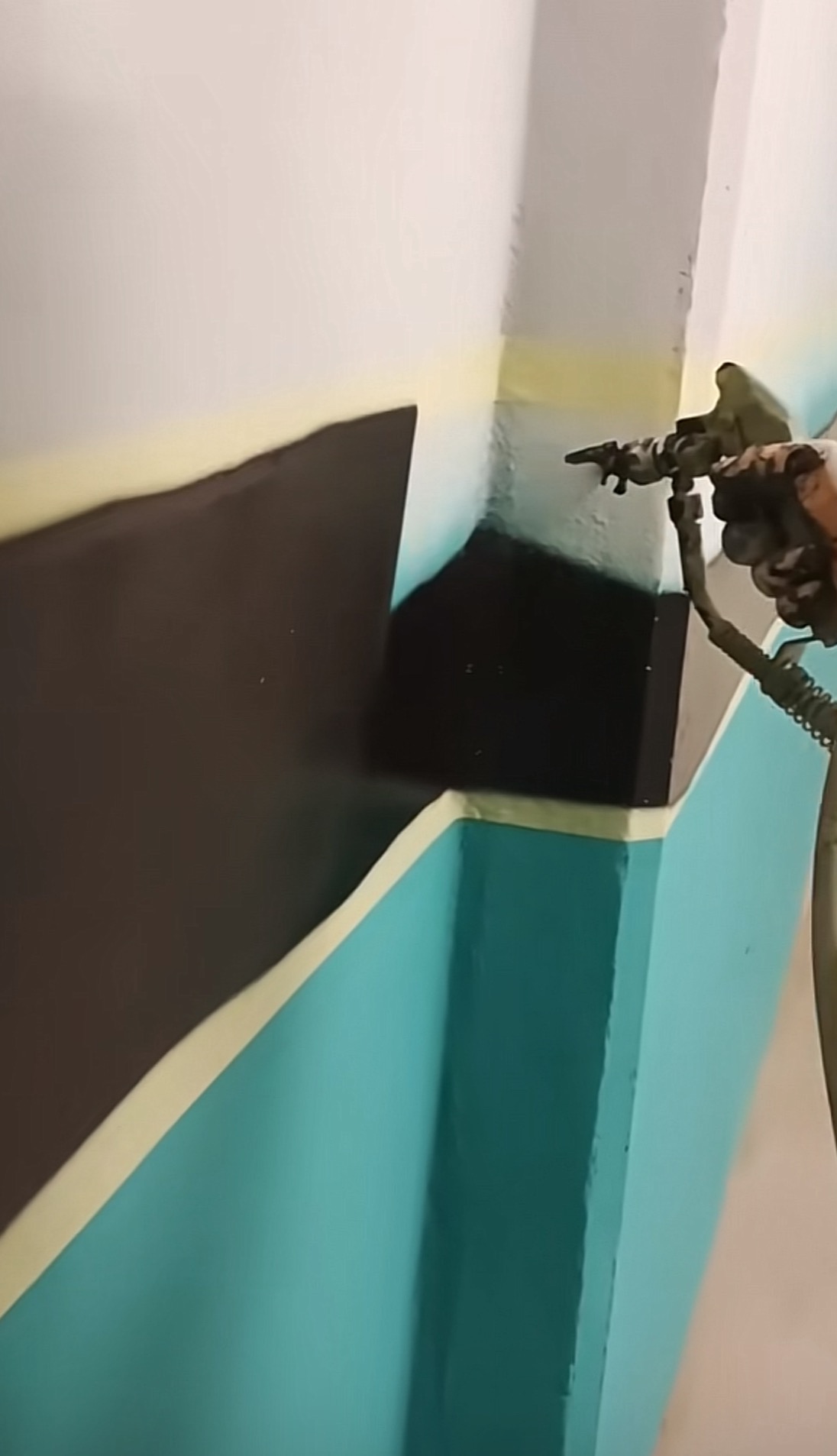 Malowanie fragmentu ściany z użyciem pistoletu natryskowego, widoczne taśmy malarskie i geometryczne wzory w kolorach turkusowym i brązowym na białym tle.