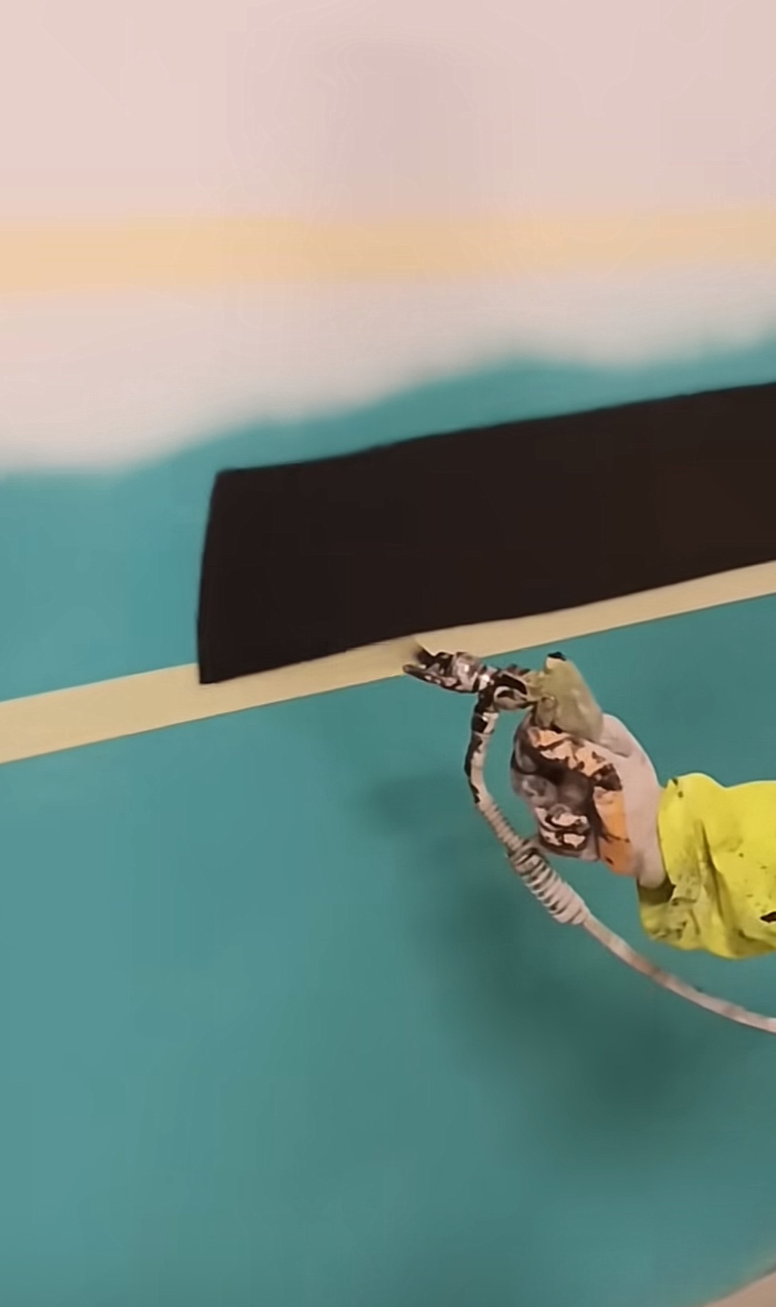 Malowanie ściany na turkusowo z czarnym pasem przy użyciu pistoletu natryskowego, zabezpieczone taśmą malarską, widoczna ręka w roboczej rękawicy i żółtej bluzie