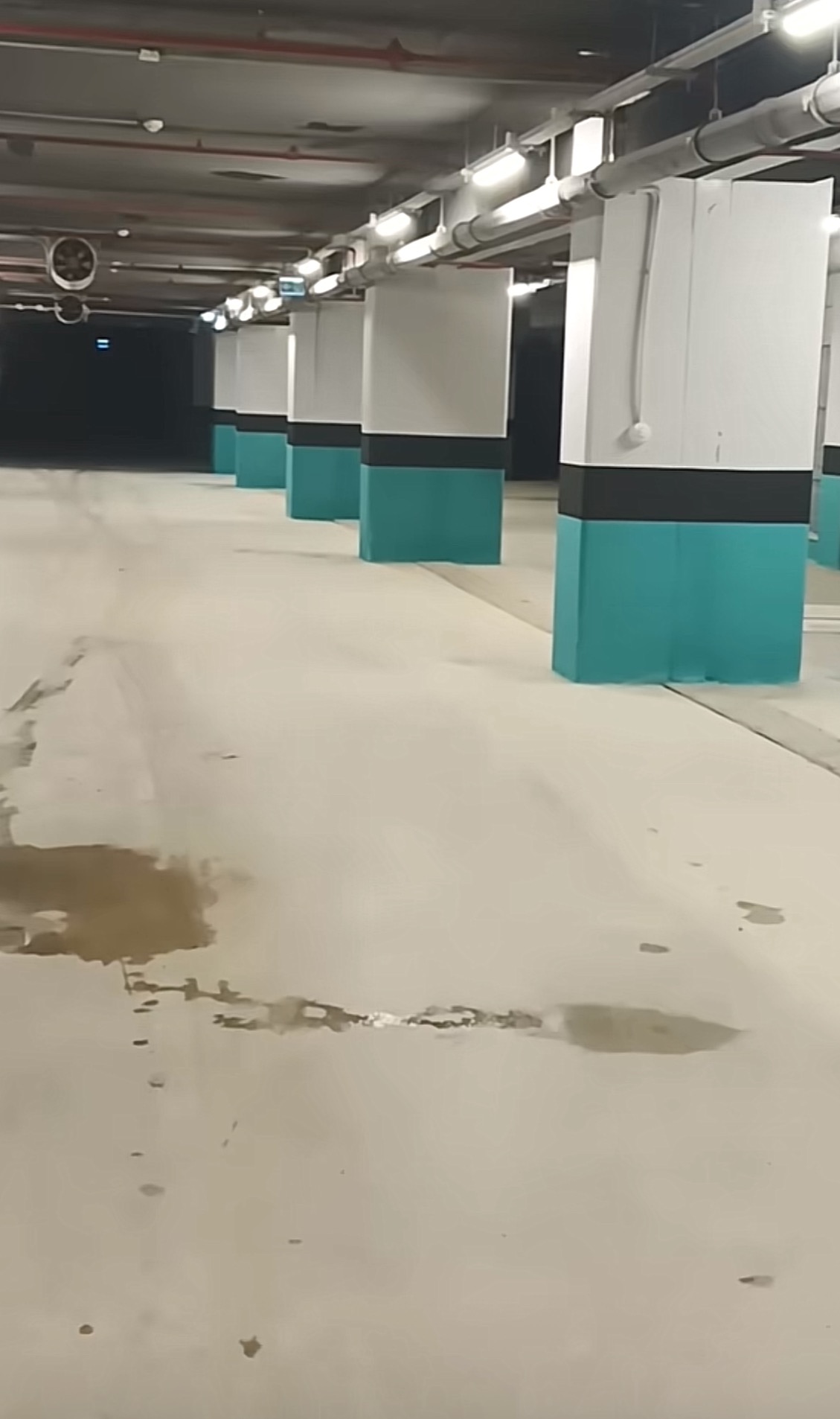 Wnętrze parkingu podziemnego z pomalowanymi na turkusowo i czarno podstawami filarów, widoczne plamy na posadzce.