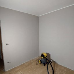 Usługi Ogólnobudowlane Paweł Soboń - Świeżo pomalowane na szaro ściany w rogu pokoju, widoczne gniazdka elektryczne bez osłon, przygotowana podłoga i odkurzacz przemysłowy Stanley SXVC20P.